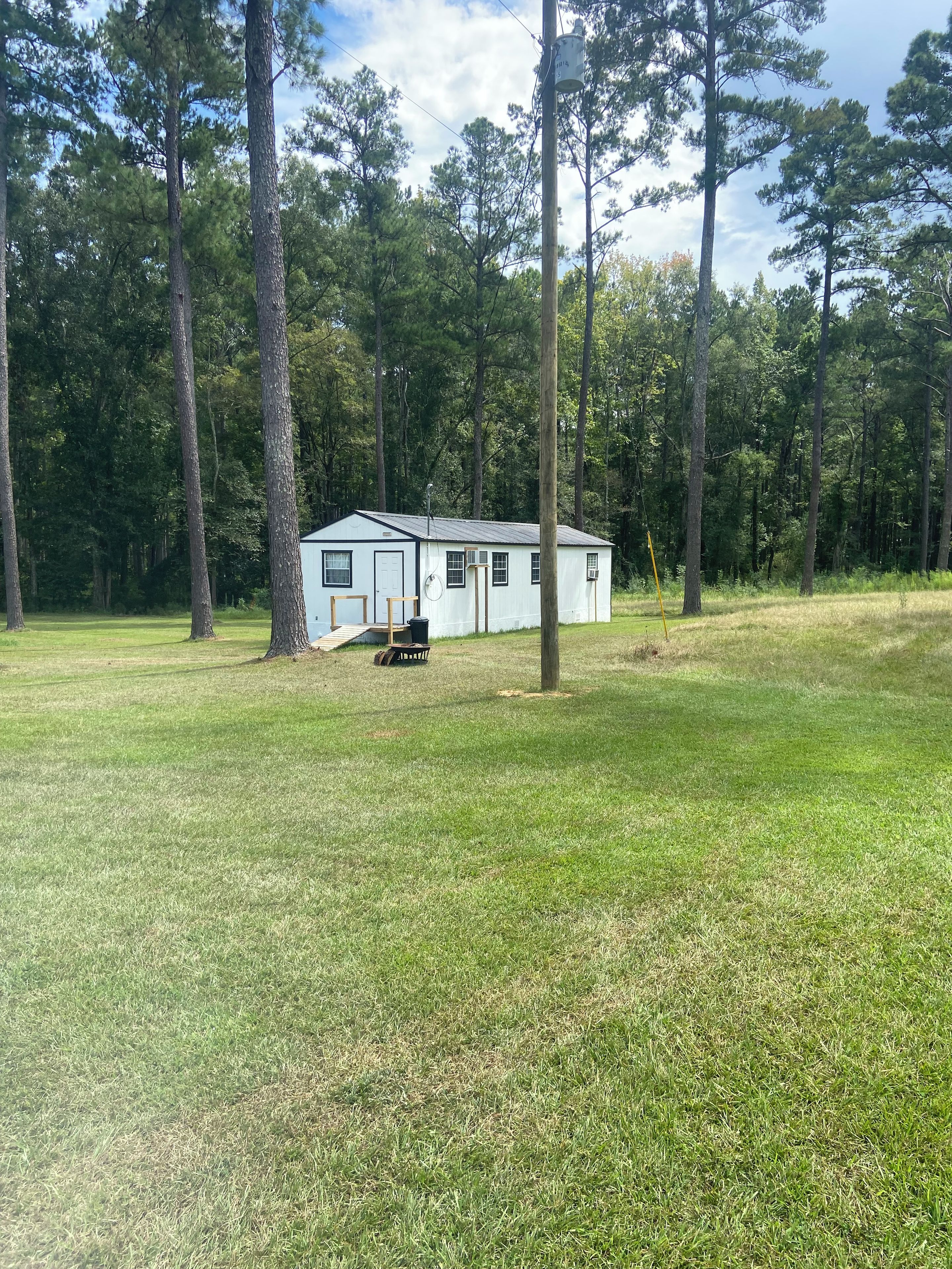 A small, single-story cabin with a front porch and a satellite dish, surrounded by a grassy field and tall trees.