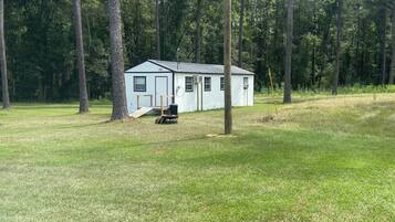 A small, single-story cabin with a front porch and a satellite dish, surrounded by a grassy field and tall trees.