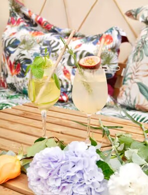 Zwei Cocktails auf einer Holztischplatte mit Blumen, im Hintergrund ein Bett mit Blumenkissen.