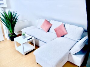 Ein modernes Wohnzimmer mit einem grauen Sofa, zwei rosafarbenen Kissen, einem kleinen weißen Beistelltisch, einer Topfpflanze und einer Heizung.