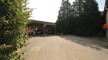 A covered parking area with a gravel surface, a car parked inside, and a building with a tiled roof in the background.