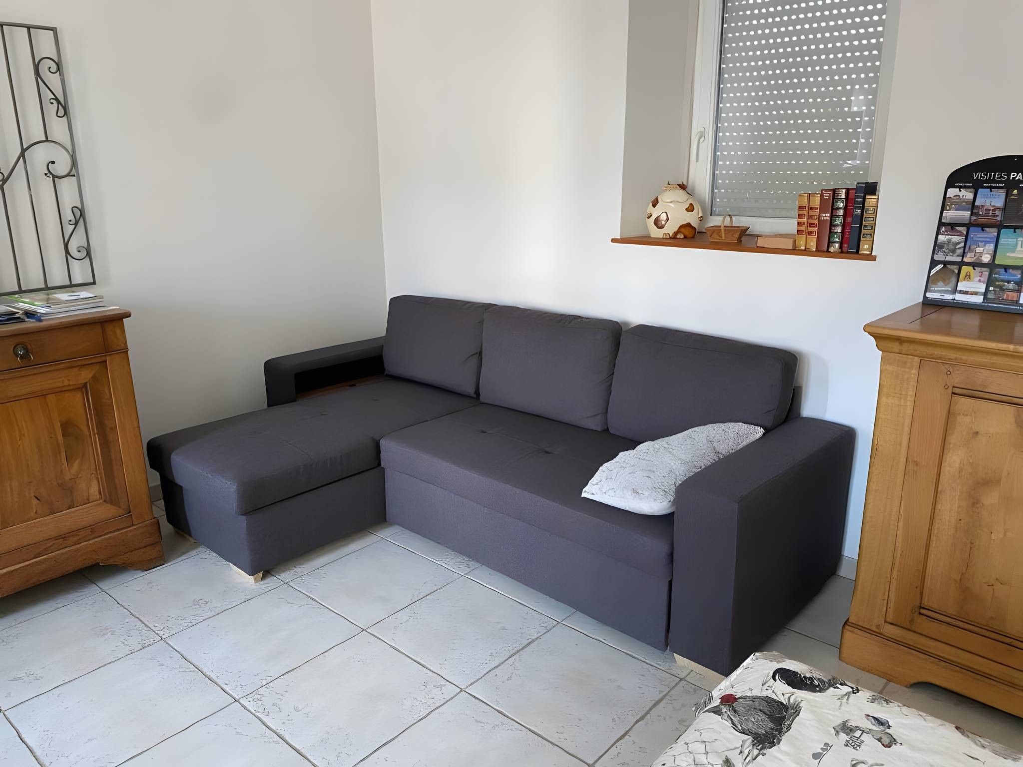 A grey sectional sofa with a white pillow, a wooden cabinet, and a window with blinds.