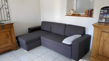 A grey sectional sofa with a white pillow, a wooden cabinet, and a window with blinds.