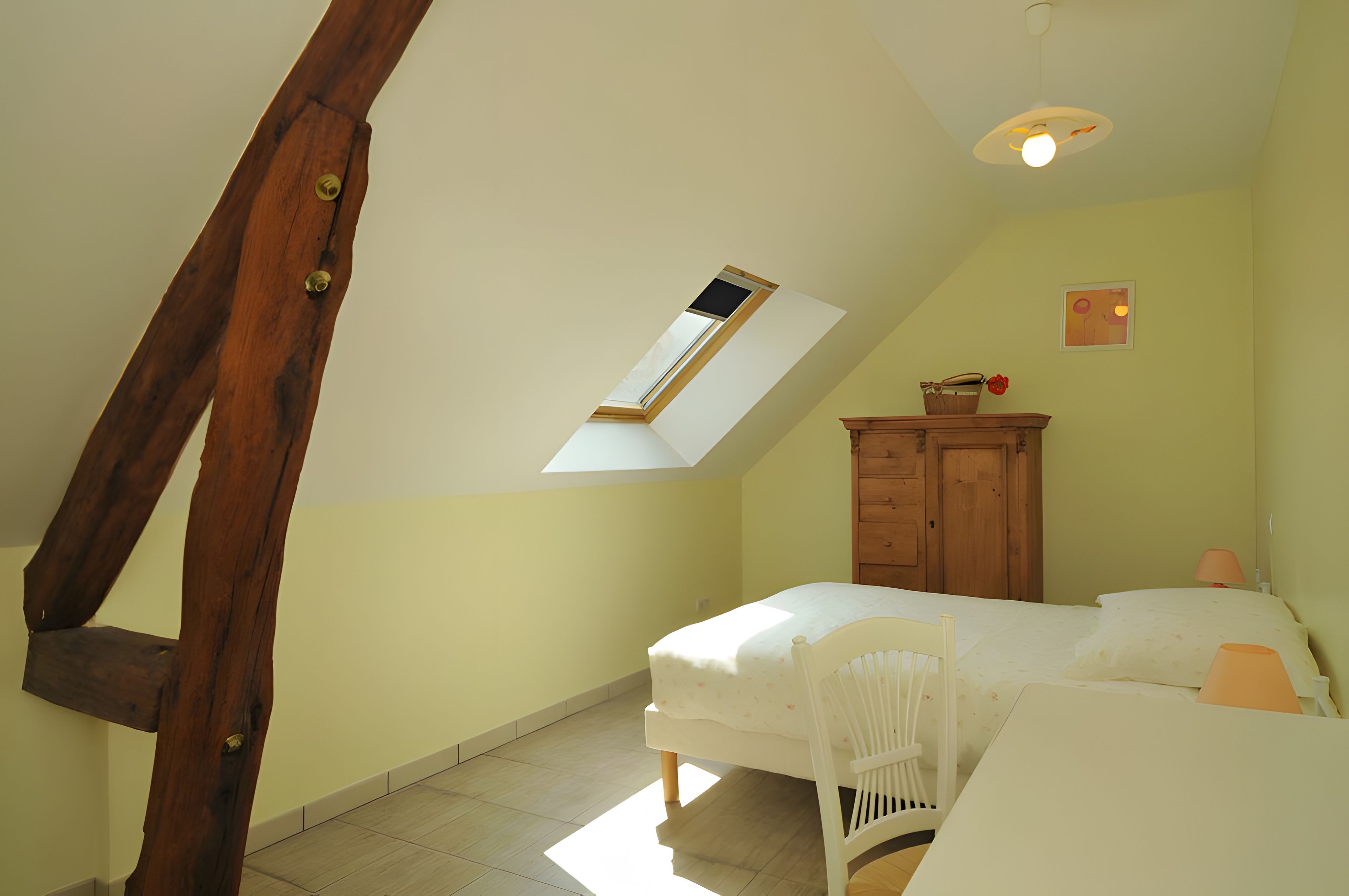 Une chambre avec un plafond en pente, un puits de lumière, une armoire en bois, un lit avec une tête de lit blanche et une petite table avec une lampe.