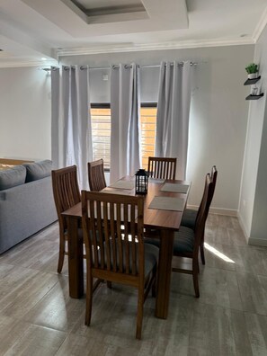 A dining area with a wooden table and chairs, a grey sofa, and a window with curtains. - Cozy 2-bedroom apartment in brilliant Lusaka with AC (Lusaka)
