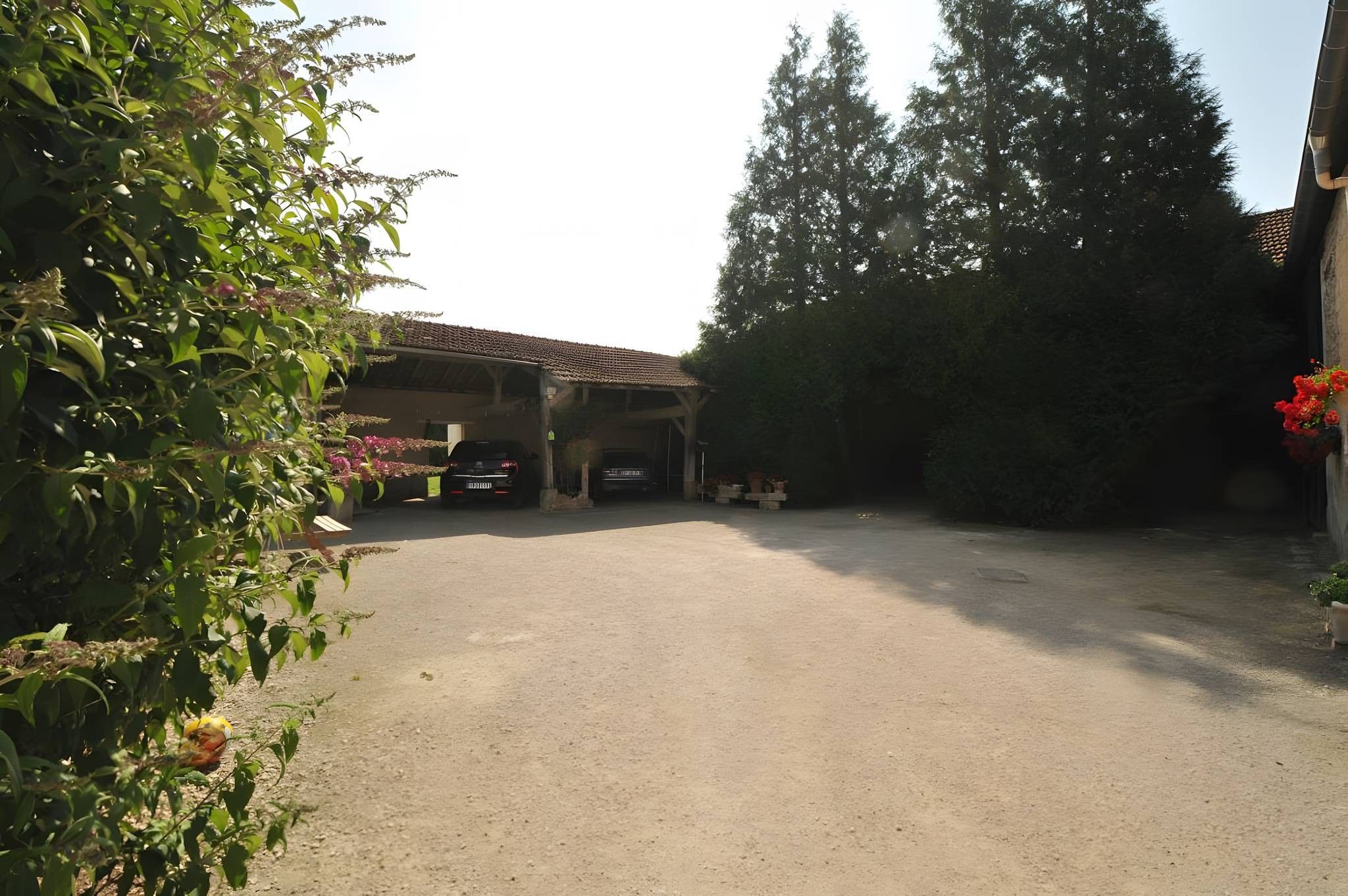 Un espace de stationnement couvert avec un sol en gravier, une voiture garée à l’intérieur, et un bâtiment avec un toit en tuiles en arrière-plan.
