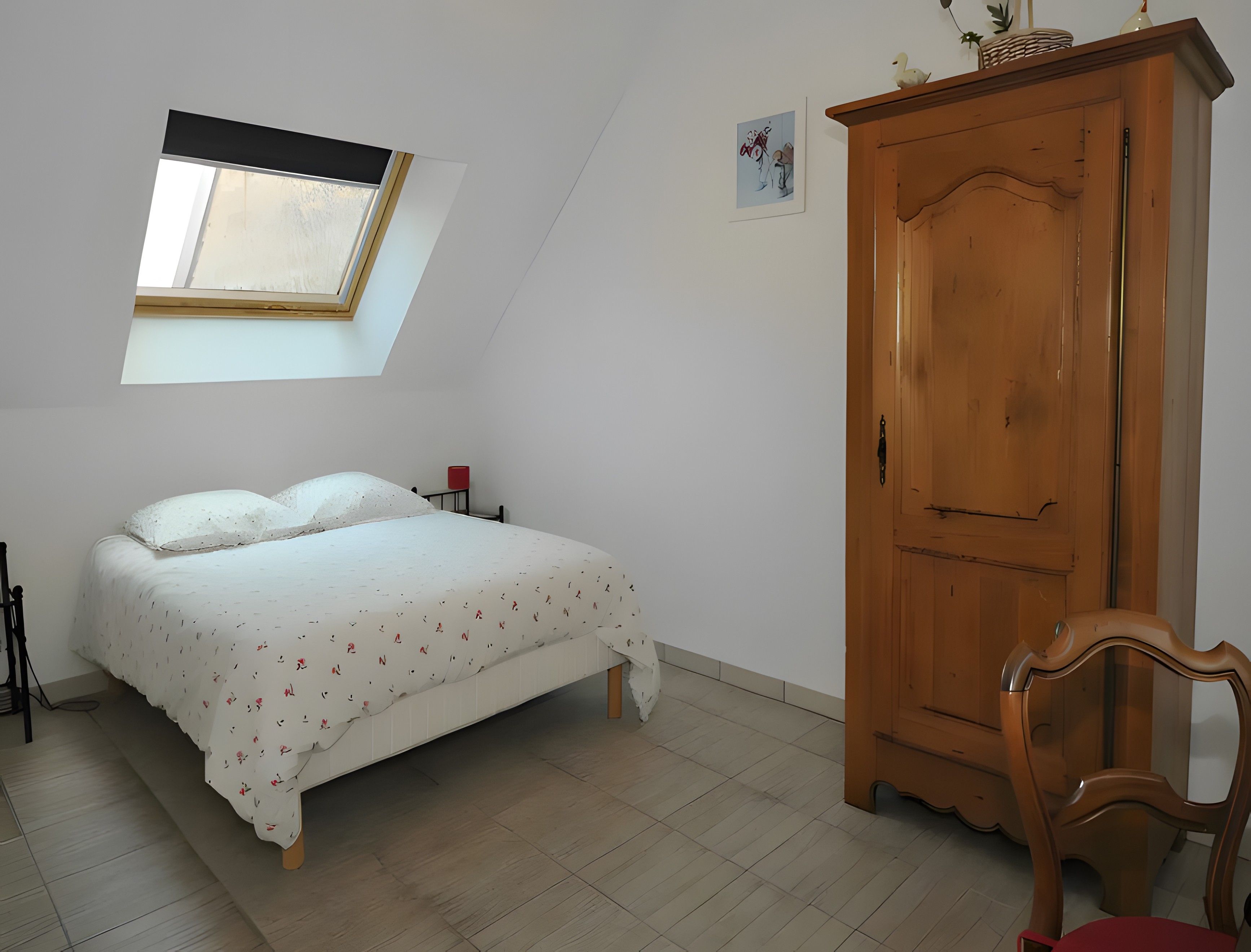 A bedroom with a bed, a wooden wardrobe, a red chair, a skylight, and a small wall decoration.