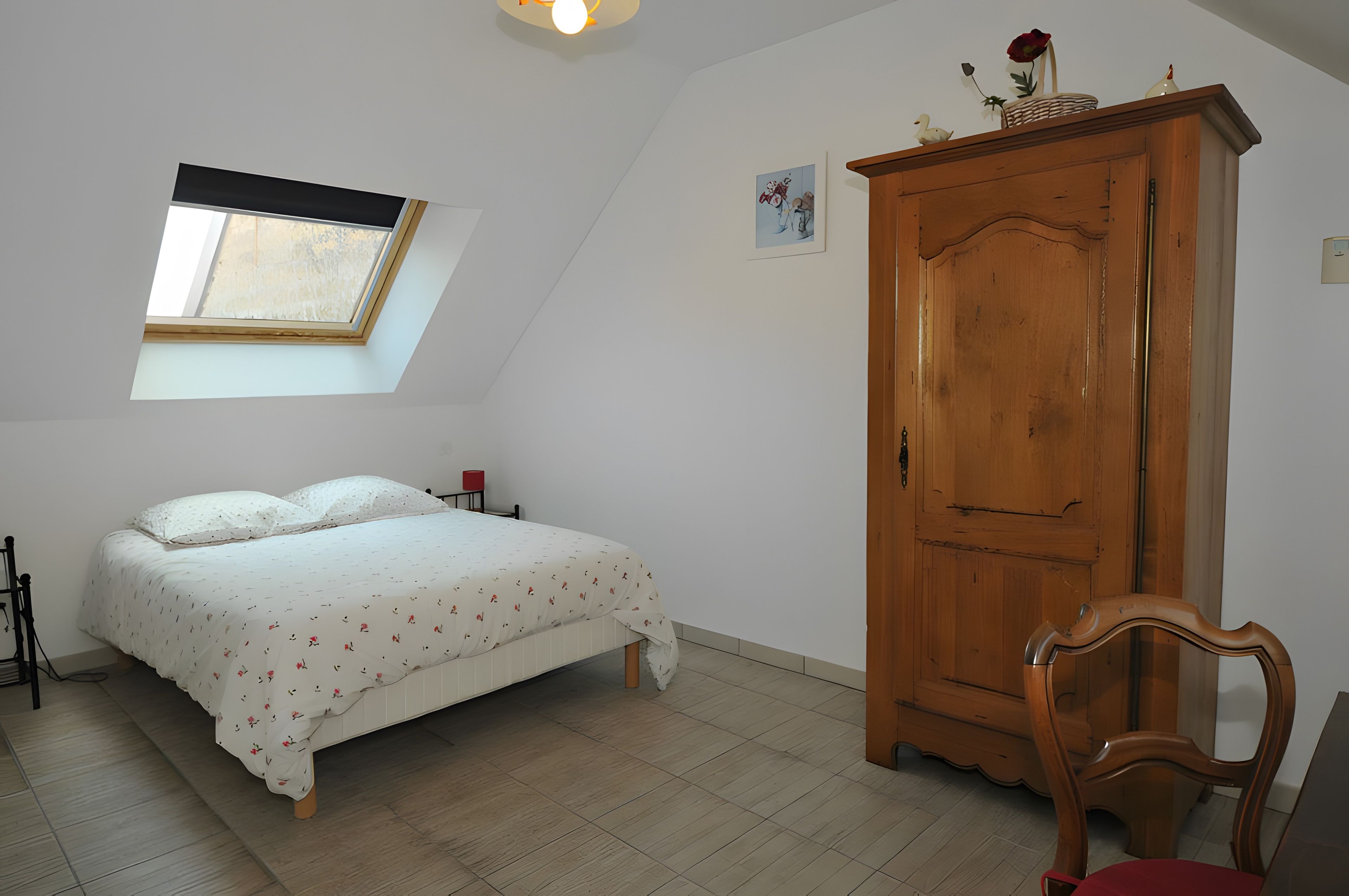 A bedroom with a bed, a wooden wardrobe, a chair, a small table, a window, and a wall-mounted light fixture.