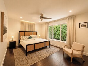 Un dormitorio con una cama, una silla, una ventana con cortinas, un ventilador de techo y una mesita de noche con lámpara.