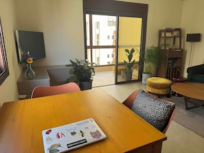 A hotel room with a wooden table, a sofa, a chair, a potted plant, a bookshelf, and a view through a glass door. - Cozy Corners- 2 bedroom apartment in amazing Byblos (byblos)