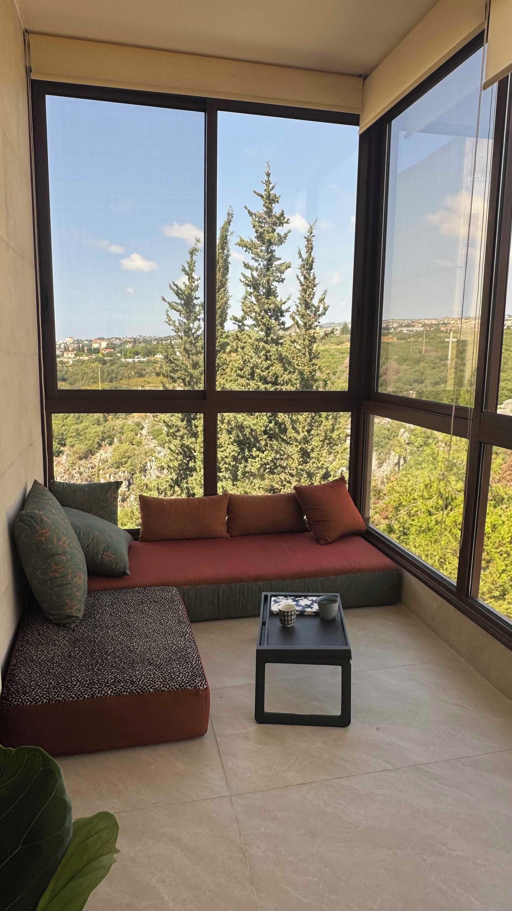 A balcony with a sofa and a small table, overlooking a cityscape with trees.