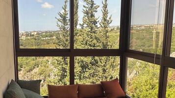 A balcony with a sofa and a small table, overlooking a cityscape with trees.