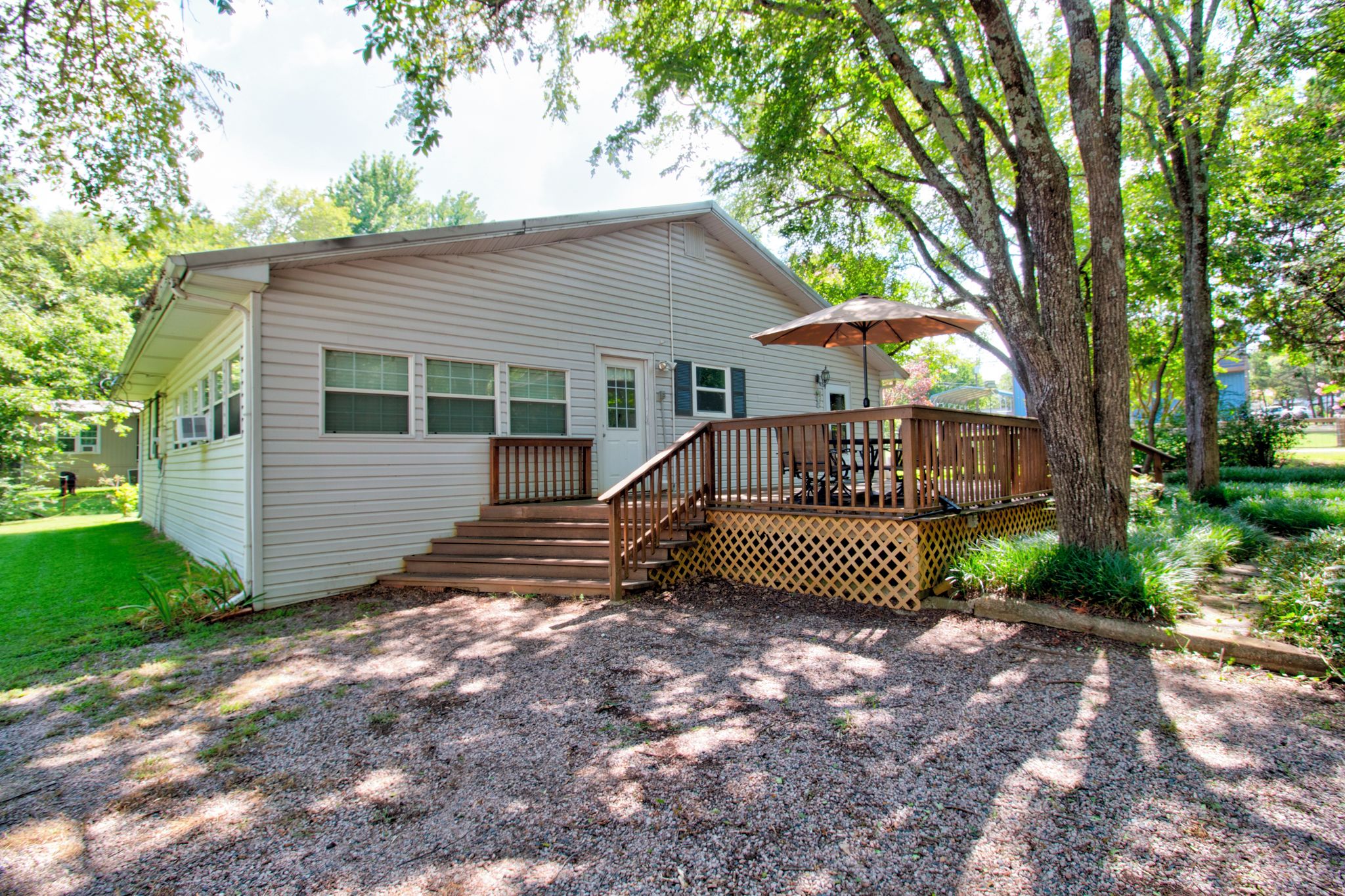 A single-story house with a deck, a staircase leading to the deck, and a gravel driveway.