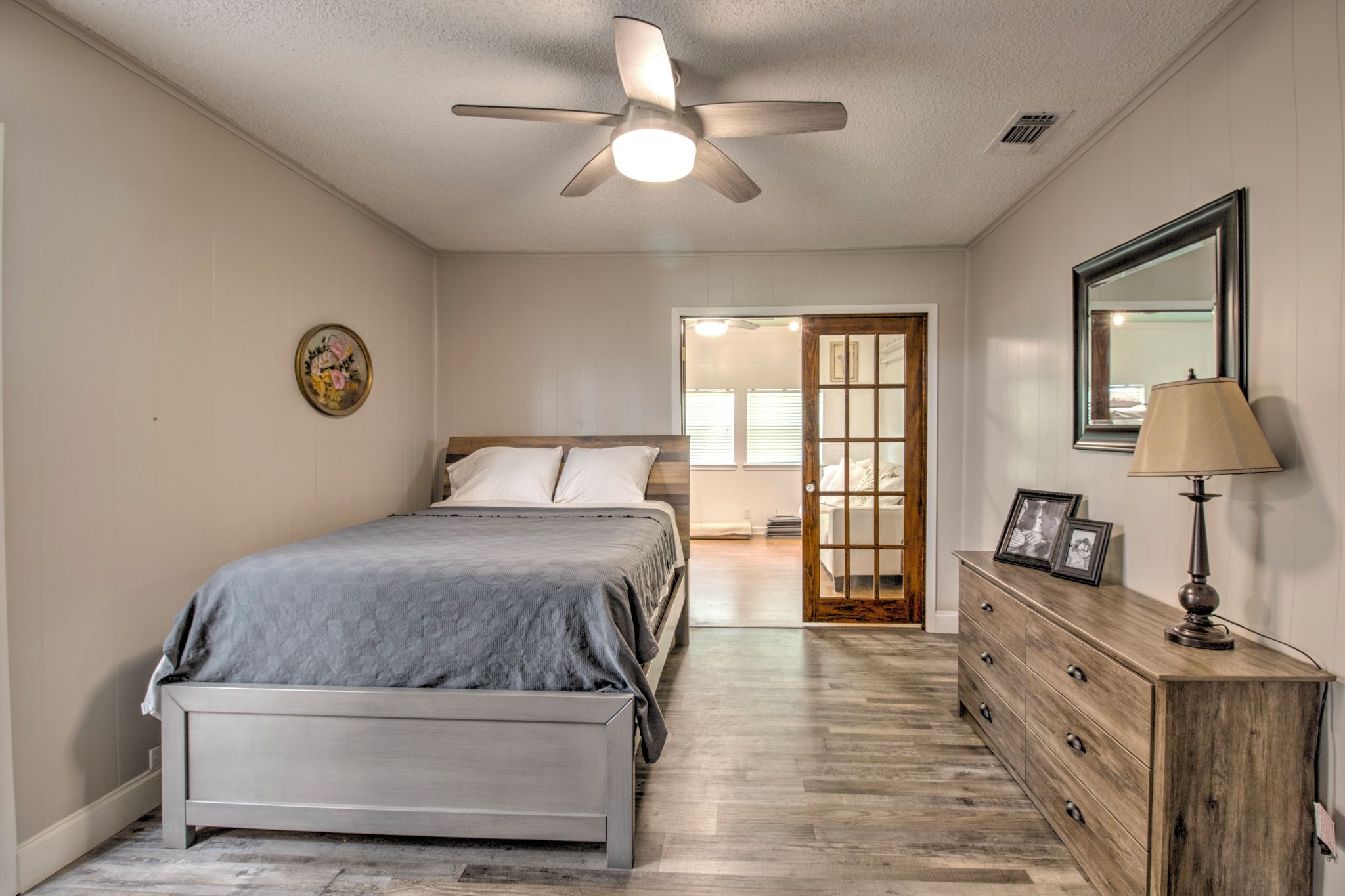 A bedroom with a bed, dresser, ceiling fan, and a door leading to another room.