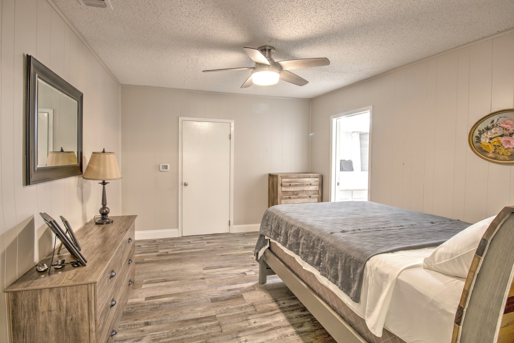 Une chambre à coucher comprenant un lit, une commode, un miroir, une lampe, un ventilateur de plafond et une porte.