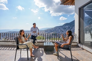 A balcony with three people seated, a table, and a view of a cityscape and mountains.