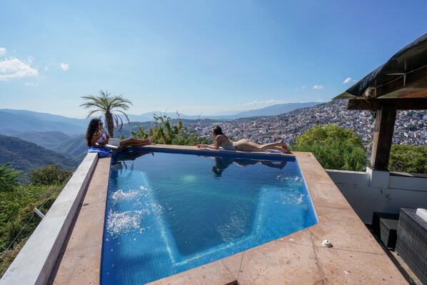An infinity pool with a city view, surrounded by greenery and a wooden deck.