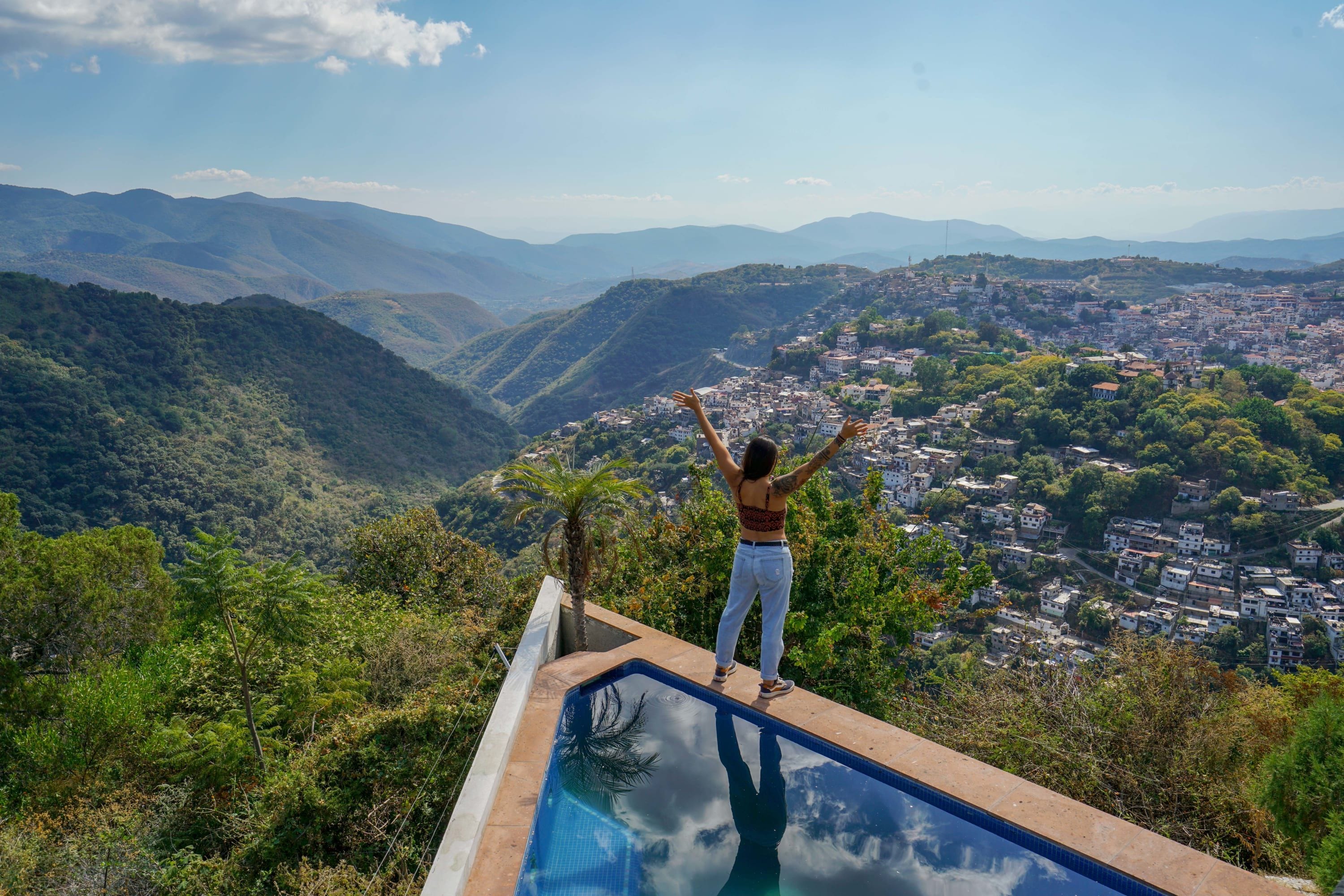 A person standing on a pool deck overlooking a mountainous landscape with a town below.