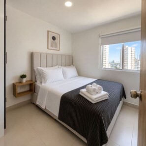 A hotel room with a bed, a small bedside table with a plant, a window with a view of buildings, and a door. - Sunlit Haze | 3BR,Beach, Sea View & Pool (Cartagena)