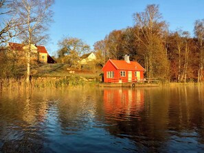 Ein rotes Haus an einem stillen See mit Spiegelungen, umgeben von Bäumen und weiteren Gebäuden.