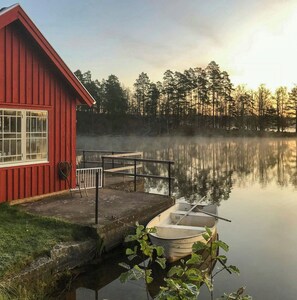 A red cabin with a dock and a small boat, surrounded by a calm lake and trees. - Lakeside cabin at Älmtasjön with sauna & hot tub (Asarum)
