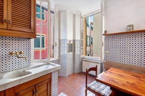 A narrow kitchen with a wooden table and chair, a sink, and a window with shutters. - Capitan Romeo Apartment (Arenzano)