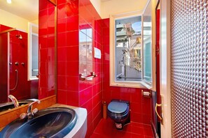 Ein Badezimmer mit roten Fliesen, einer Badewanne, einer Toilette, einem Fenster mit Blick auf Gebäude und einer Duschkabine.