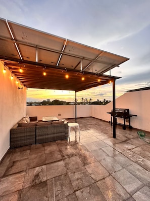 Um pátio externo com área de estar coberta, churrasqueira e vista para o pôr do sol.