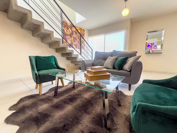 Uma sala de estar moderna com uma mesa de centro de vidro, duas poltronas verdes, um sofá cinzento e uma escada com um mural colorido.