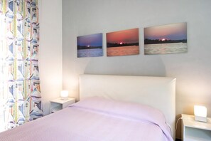 A bedroom with a bed, two bedside tables with lamps, a patterned curtain, and three wall-mounted sunset paintings. - Casa Andrea - Two Bedroom Apartment, Sleeps 4 (Arenzano)
