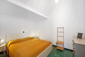 Quarto com cama, mesa de cabeceira com luminária, escada de madeira e um pequeno armário.