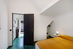 Una habitación con una cama, una puerta, una mesita con lámpara y una escalera.