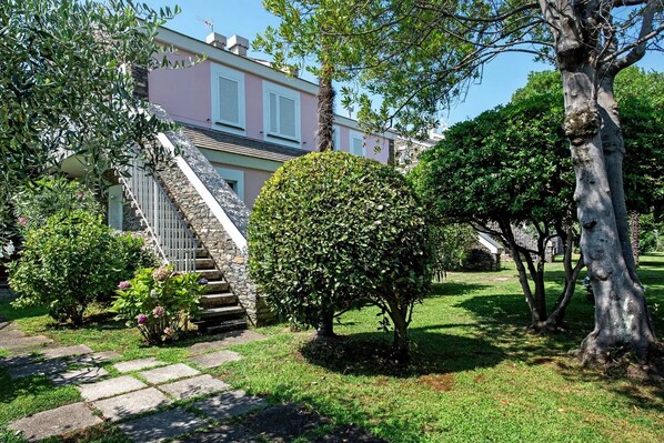 Escadaria de pedra que leva a um edifício com fachada rosa e persianas brancas, cercado por vegetação bem cuidada e céu claro.