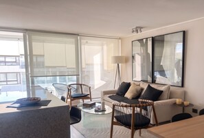 Ein modernes Wohnzimmer mit einem Glastisch, einer Couch und einem Sessel. Es gibt ein großes Fenster mit Jalousien, eine Stehlampe und ein gerahmtes Kunstwerk an der Wand.