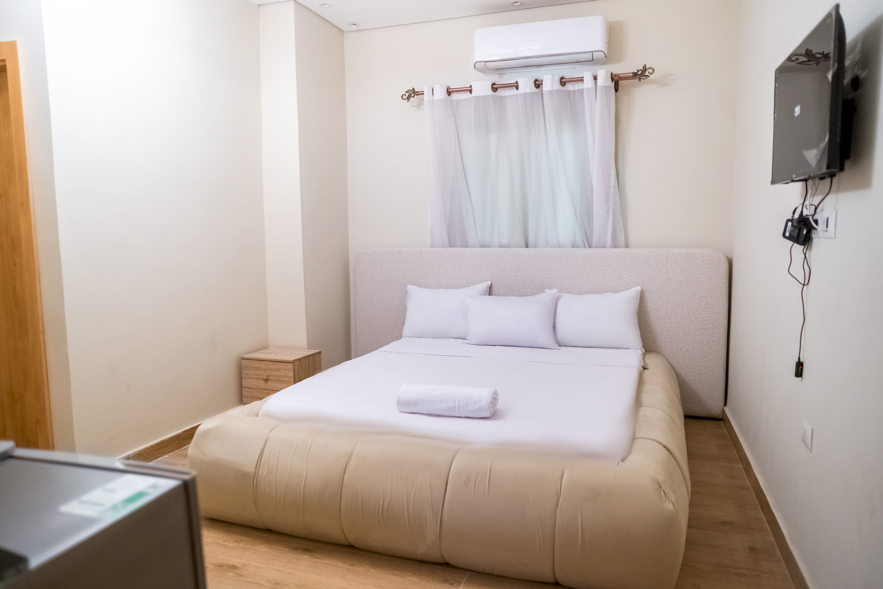 A neatly made bed with white linens and a rolled towel, a small bedside table, and a wall-mounted TV.
