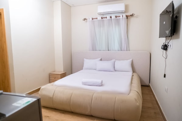 A neatly made bed with white linens and a rolled towel, a small bedside table, and a wall-mounted TV.