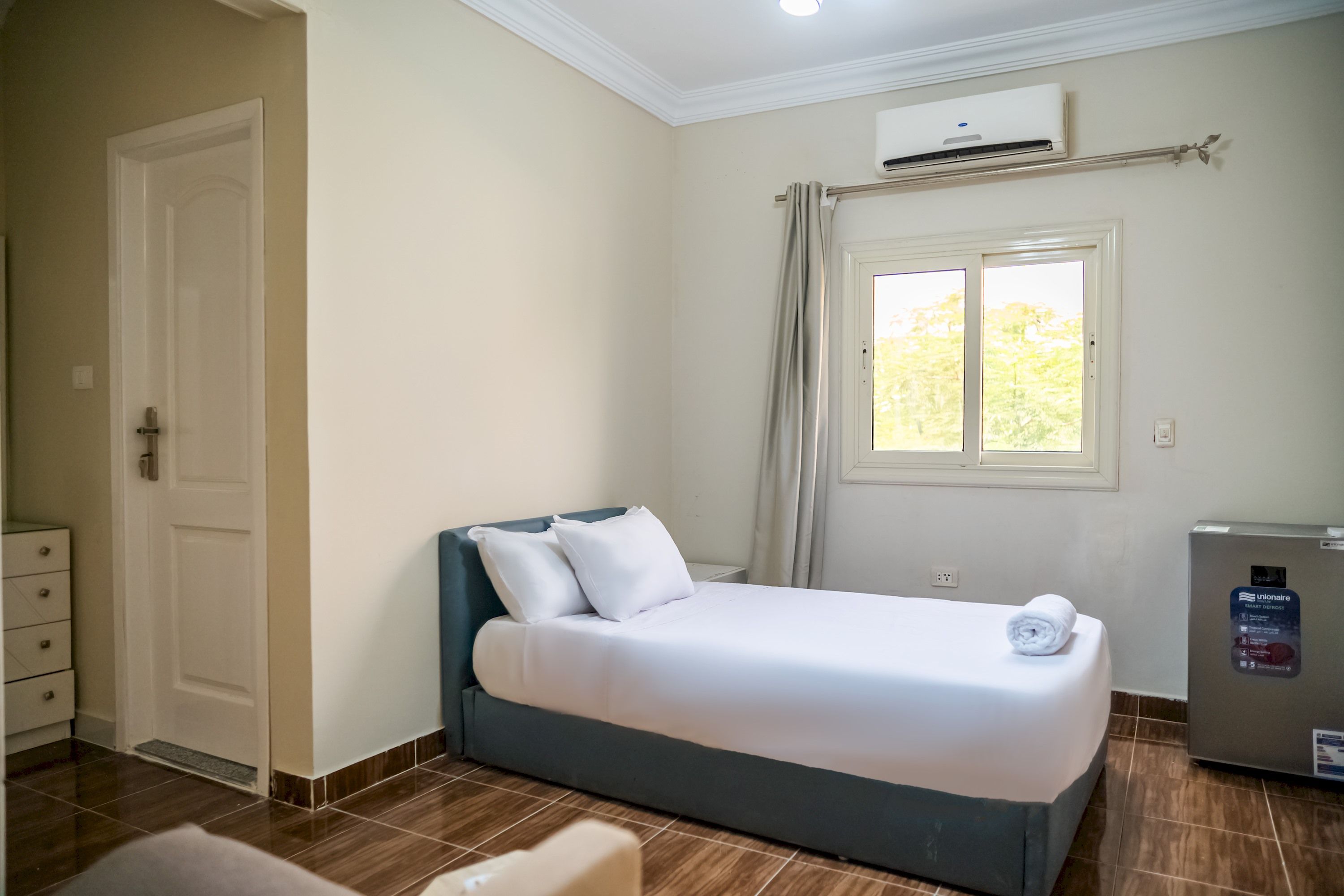 Una habitación de hotel con cama, ventana con cortinas, unidad de aire acondicionado y un refrigerador pequeño.