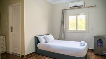 Una habitación de hotel con cama, ventana con cortinas, unidad de aire acondicionado y un refrigerador pequeño.