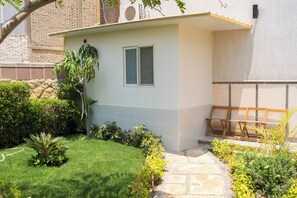 A small building with a window, surrounded by a well-maintained garden with various plants and a stone pathway. - Single Room with Private Bathroom (Sheikh Zayed City)