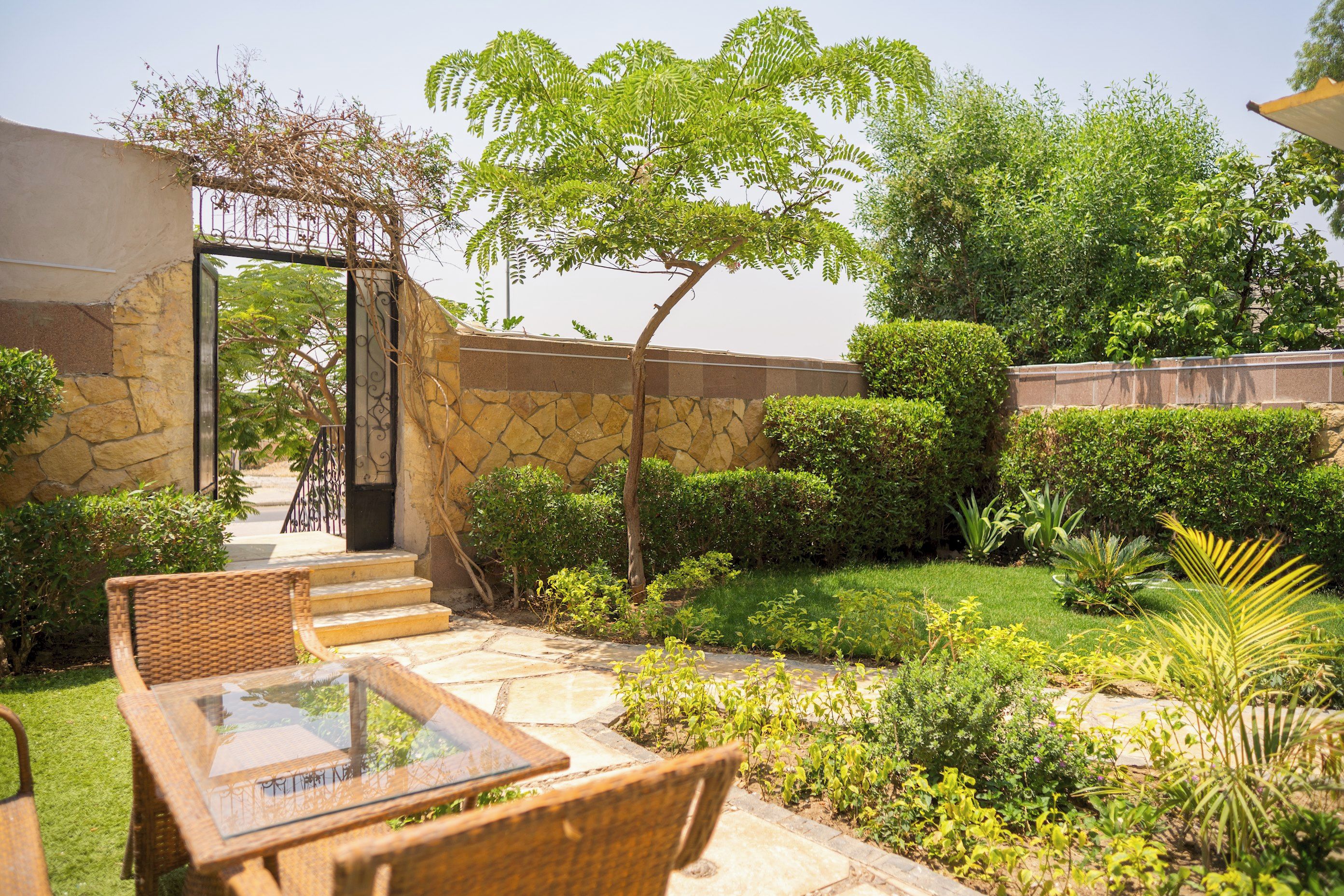 Un jardín con pared de piedra, una pérgola y una mesa de vidrio con sillas de mimbre.