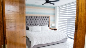 Une chambre à coucher avec une tête de lit capitonnée, un lit avec une literie blanche, une table de chevet avec une lampe et une fenêtre avec des stores.