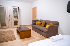 A room with a wooden wardrobe, a brown sofa with cushions, a small wooden table, and a white bed with a rolled-up white towel. - Double Room with Private Bathroom (Sheikh Zayed City)