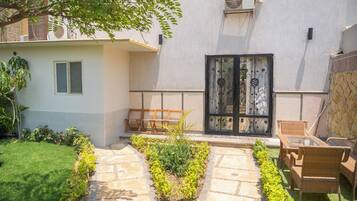 Un patio con un camino de piedra, un banco de madera, una silla de mimbre y una puerta con detalles de metal ornamentado.