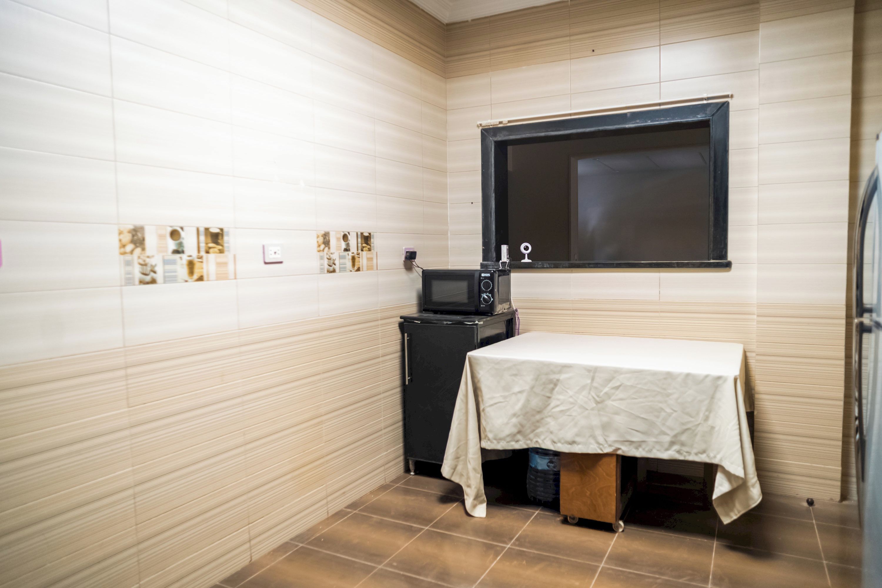 Pequeña habitación con suelo de baldosas, una mesa cubierta con tela blanca, un televisor negro y una ventana con marco negro.