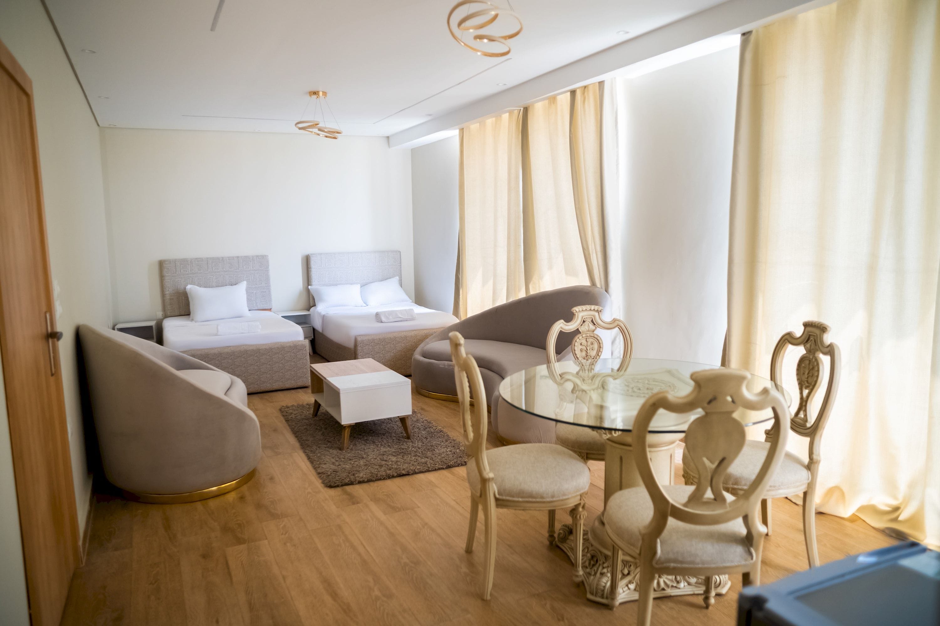 A modern hotel room with a sofa, two beds, a dining table with chairs, and a large window with curtains.