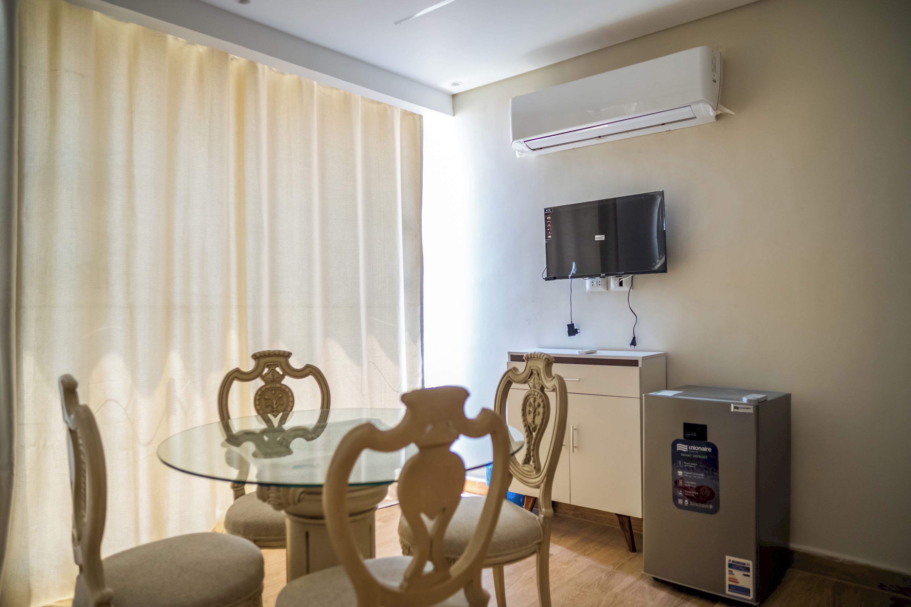 Habitación de hotel con una mesa de vidrio, dos sillas, un televisor montado en la pared y un minibar.