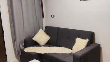 A living room with a black sofa, two white fluffy pillows, a white furry ottoman, a patterned rug, a wall-mounted air conditioner, and a framed artwork.