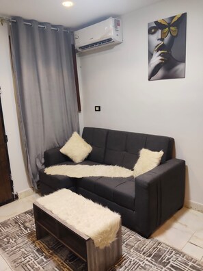 Un salón con un sofá negro, dos almohadillas blancas y mullidas, un otoman blanco y peludo, una alfombra estampada, un aire acondicionado de pared y una obra de arte enmarcada.