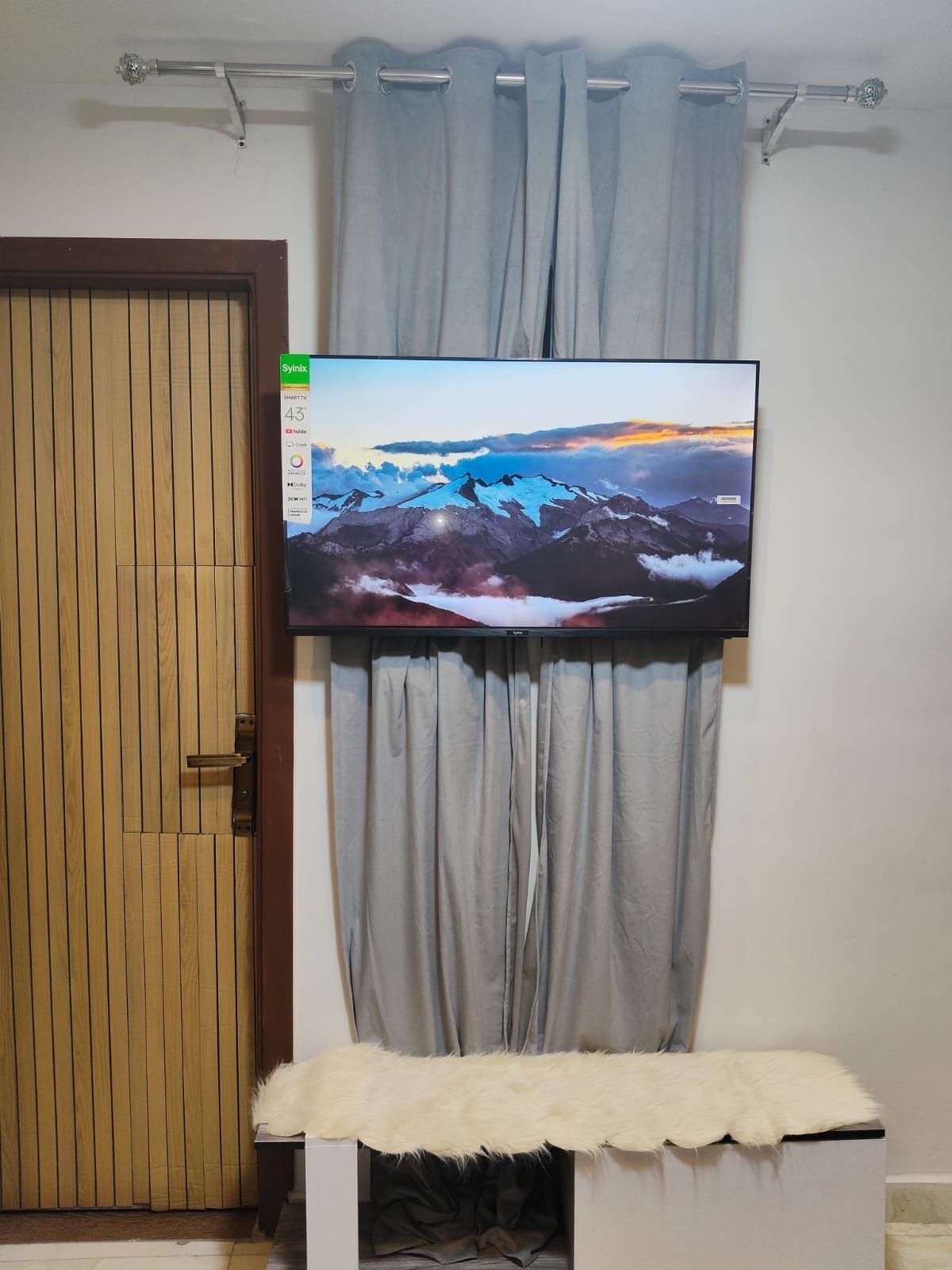 A flat-screen TV mounted on a wall above a grey curtain, with a wooden door to the left.