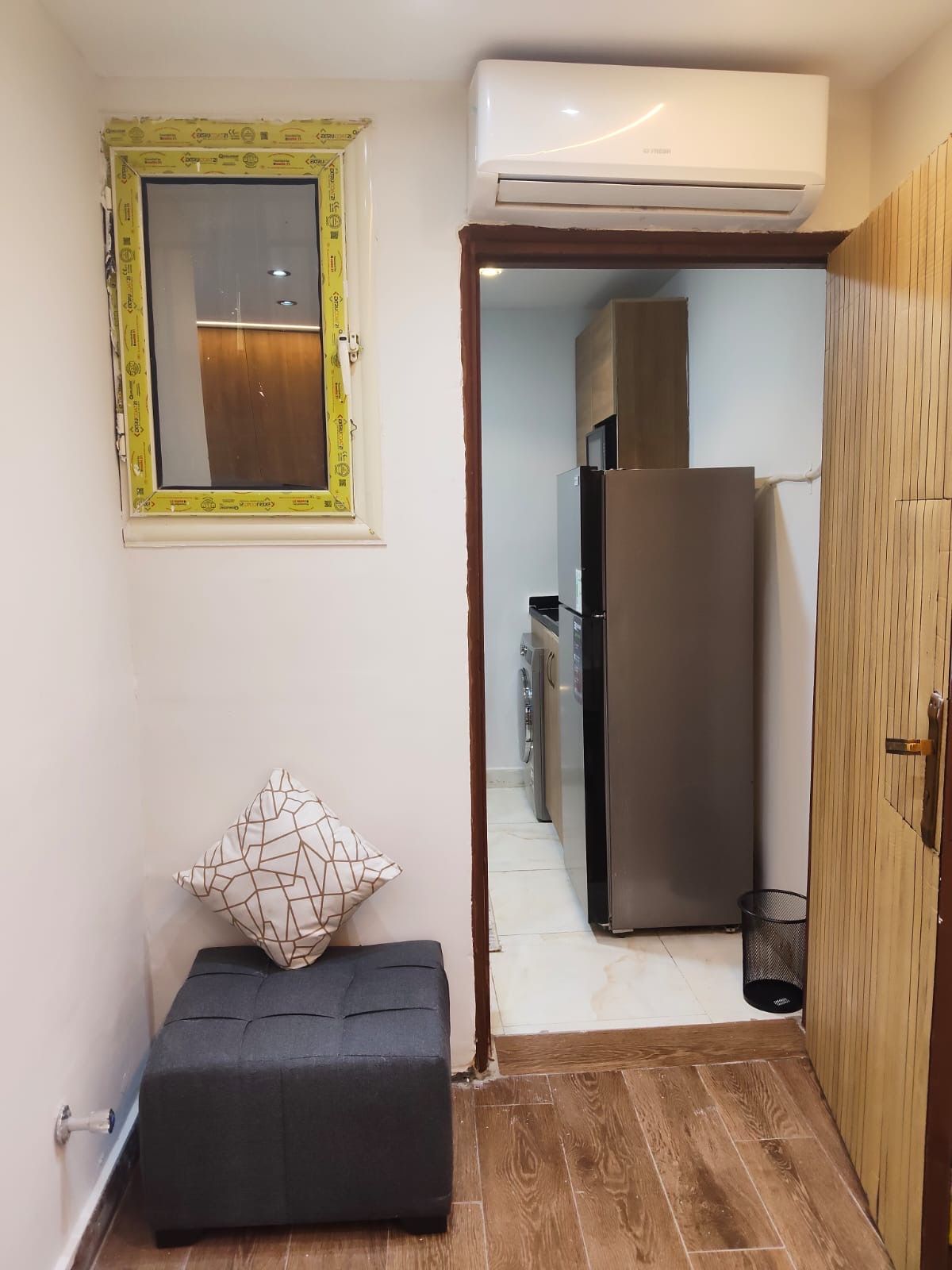 Eine kompakte Küchenzeile mit Kühlschrank, einem Fenster mit gelbem Rahmen und einer kleinen Sitzecke mit Kissen.